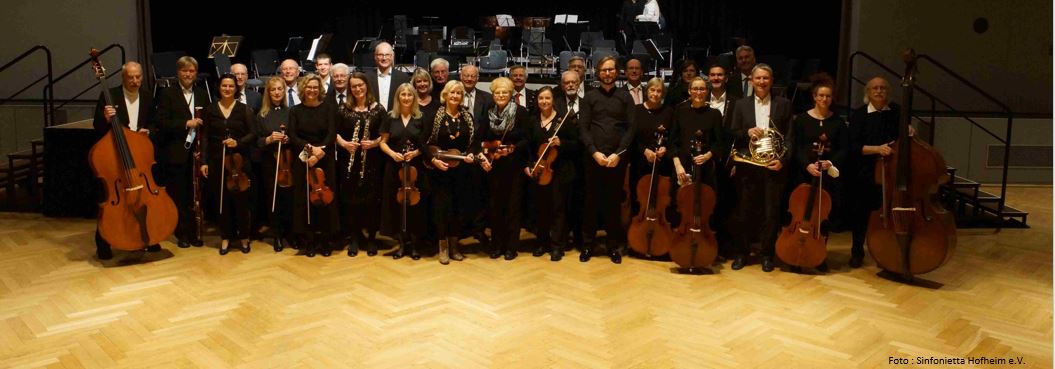 Gruppenbild der Sinfonietta Hofheim. Das Ensemble steht mit den Instrumenten vor der Bühne der Stadthallte Hofheim.
